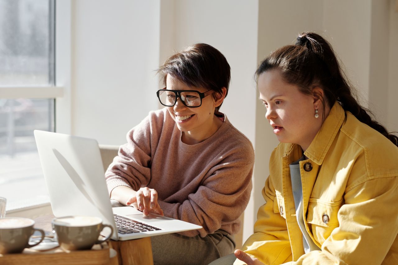 Two women using a laptop in a sunlit room, enjoying each other's company.
