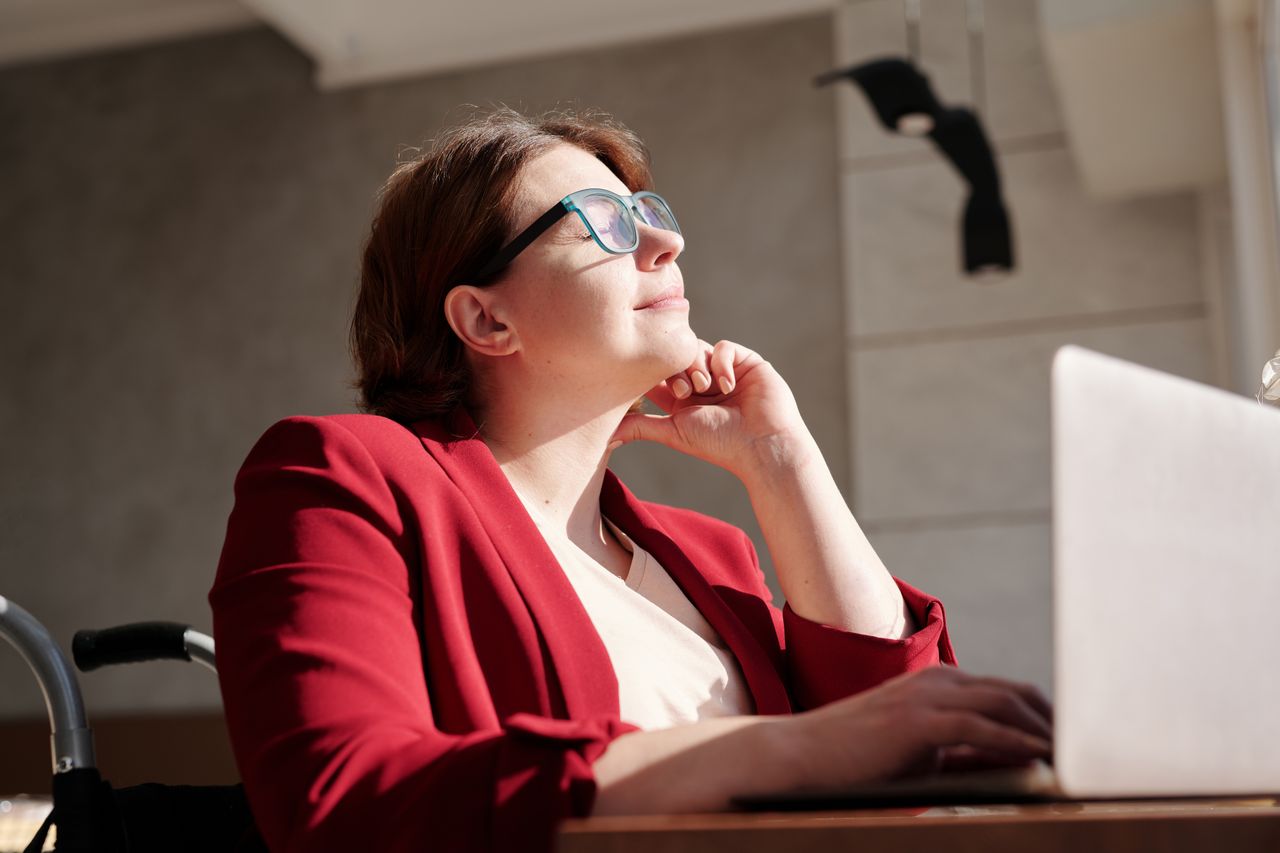 Woman in wheelchair wearing red suit and glasses, working on a laptop in a bright café.