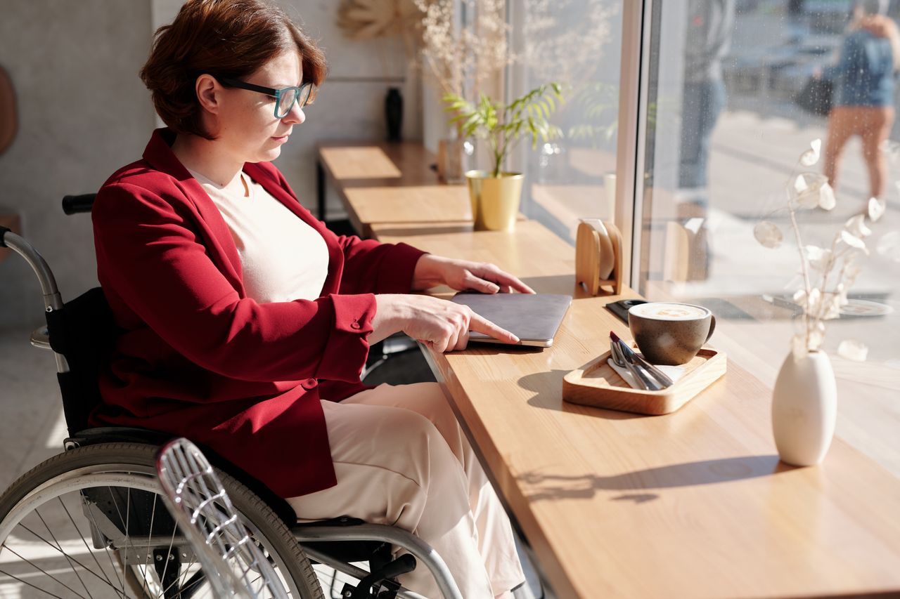 A woman in a wheelchair uses a tablet in a sunlit café setting, enjoying a morning coffee.