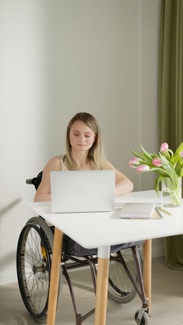 A woman in a wheelchair working on a laptop at a home office desk with flowers.