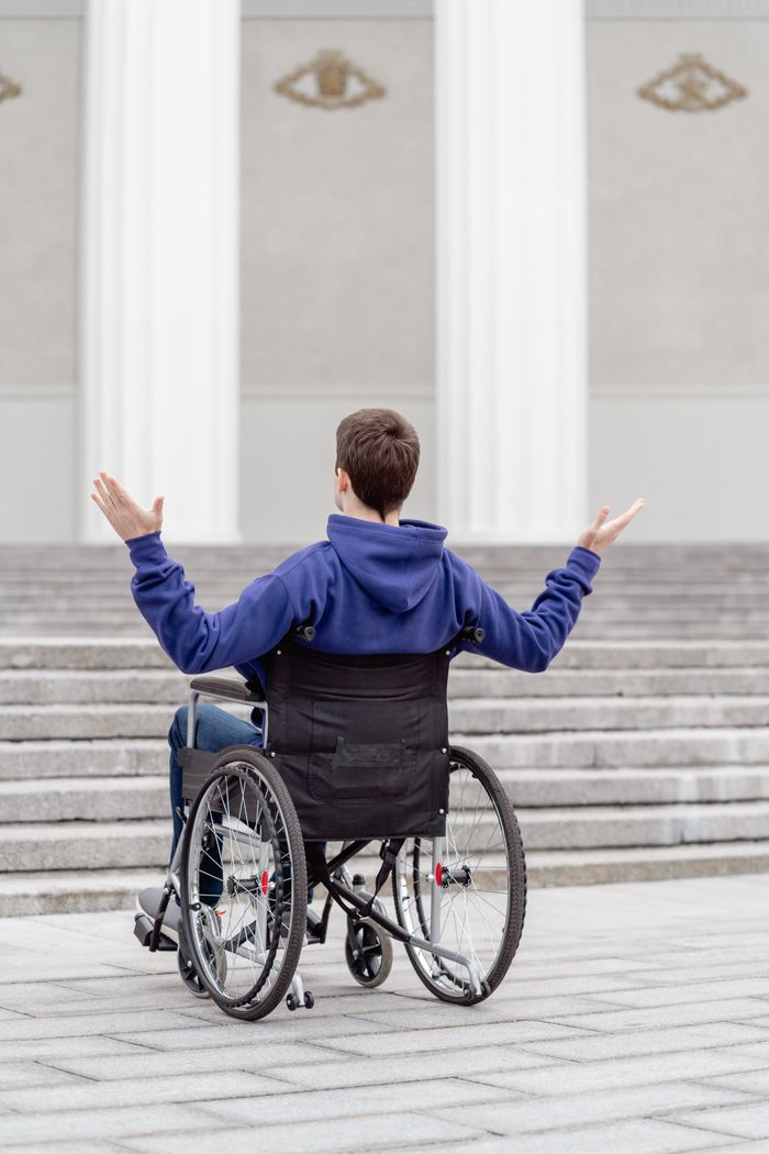 our-experience Young man in a wheelchair facing stairs, arms raised, outdoors.