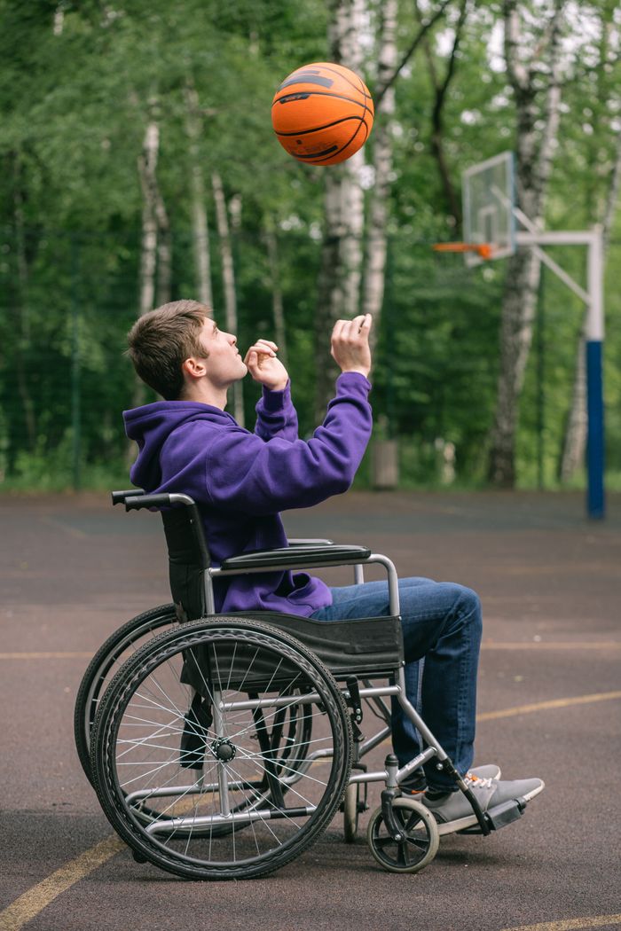 A young man in a purple hoodie plays basketball from his wheelchair on an outdoor court.